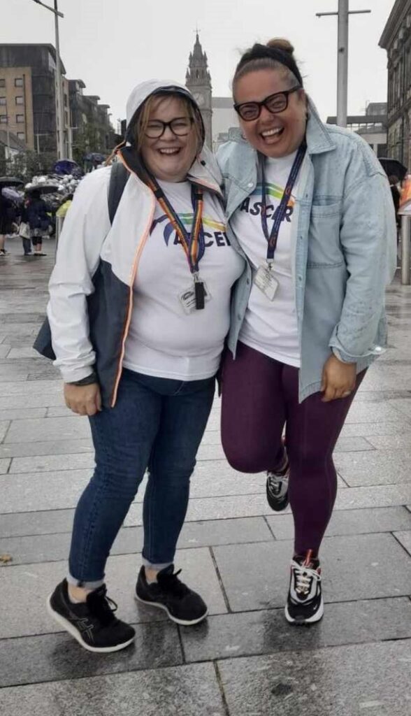 Two women stand smiling in the rain. They are wearing white t-shirts with ASCERT's logo in Pride colours.